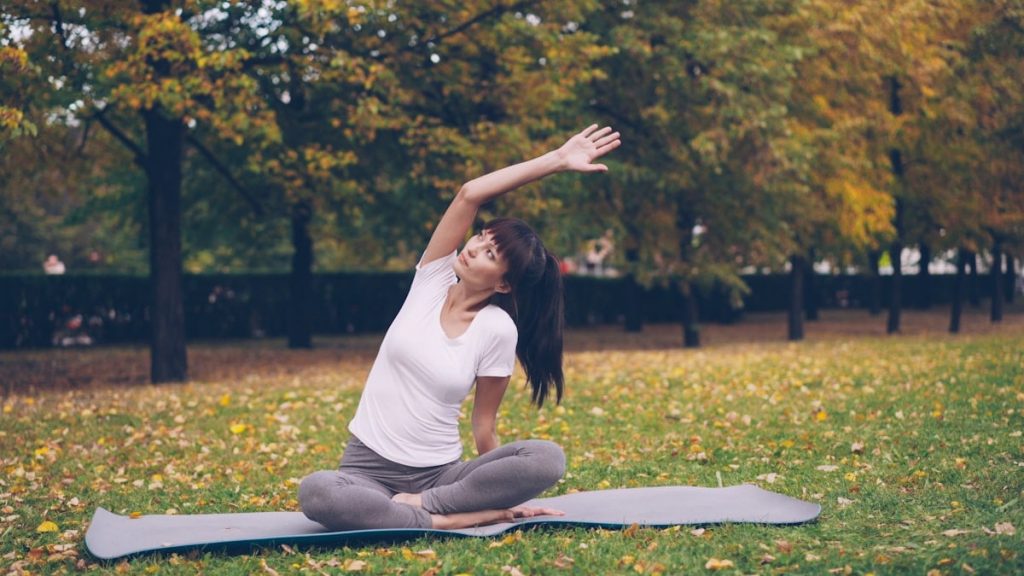 Beautiful girl is concentrated on yoga practice sitting on mat in park in lotus position and bending sideways raising arm. Healthy lifestyle, sports and recreation concept.