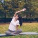 Beautiful girl is concentrated on yoga practice sitting on mat in park in lotus position and bending sideways raising arm. Healthy lifestyle, sports and recreation concept.