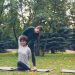 Diligent yoga student is learning Cow Face position sitting on mat and stretching arms while experienced teacher is helping her, girls are talking and laughing at outdoor ptactice.